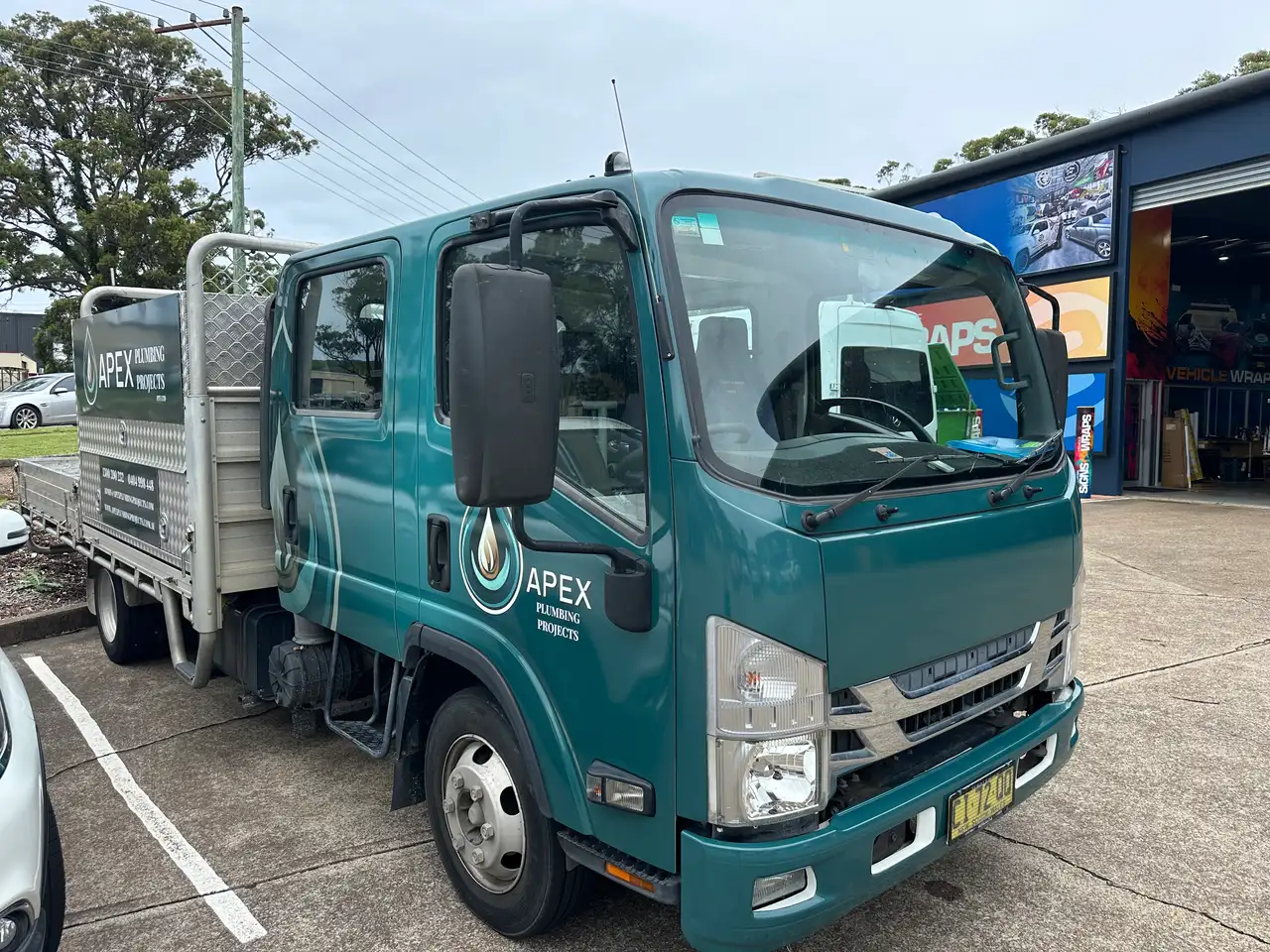 Teal Isuzu crew-cab truck with an aluminium tray, the Apex Plumbing Projects water-drop logo on the door and the contact details on the tray box, parked at the Big Colour workshop