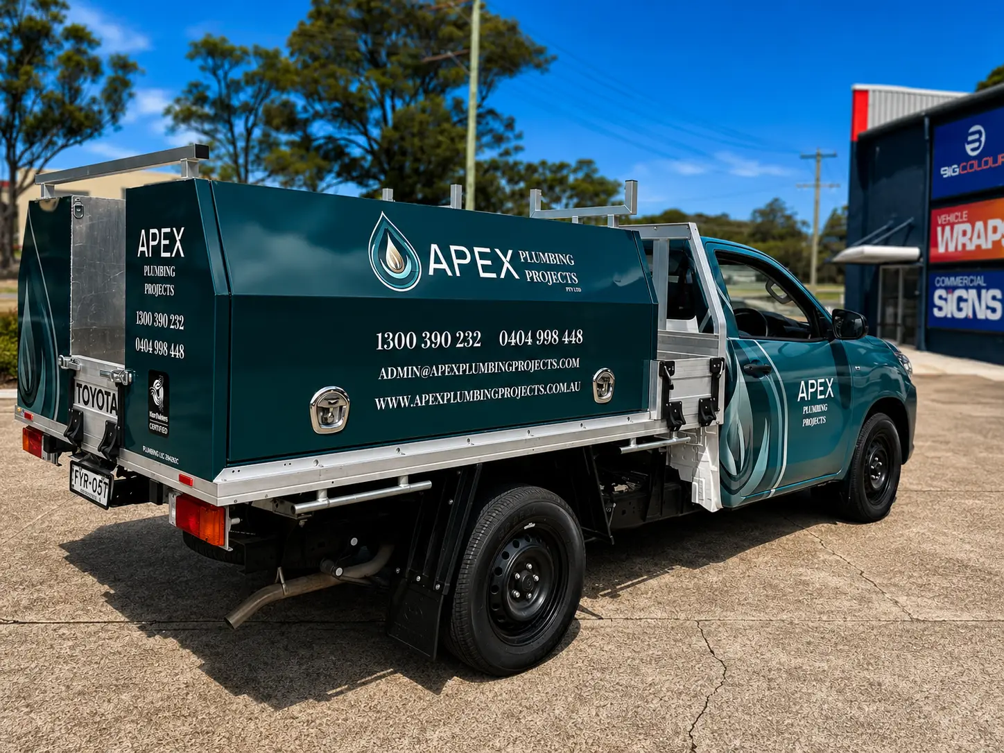 Deep teal Toyota single-cab tray ute, rear three-quarter view, with an Apex Plumbing Projects toolbox canopy carrying the water-drop logo, phone numbers and website, parked outside the Big Colour workshop