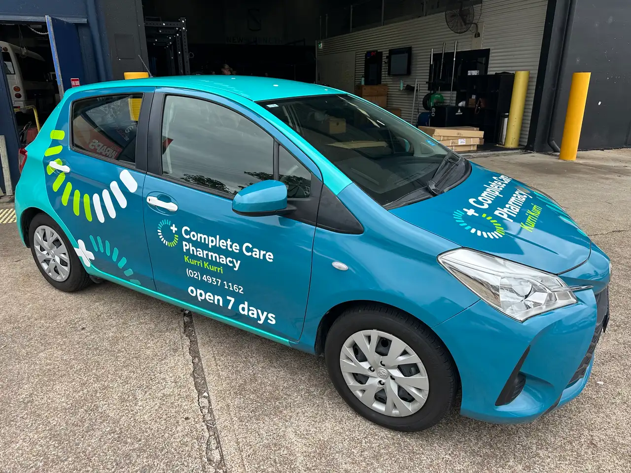 The Complete Care Pharmacy delivery car, a turquoise wrapped Toyota Yaris hatchback, seen from the front three-quarter with the Complete Care Pharmacy Kurri Kurri logo, (02) 4937 1162 and open 7 days along the side