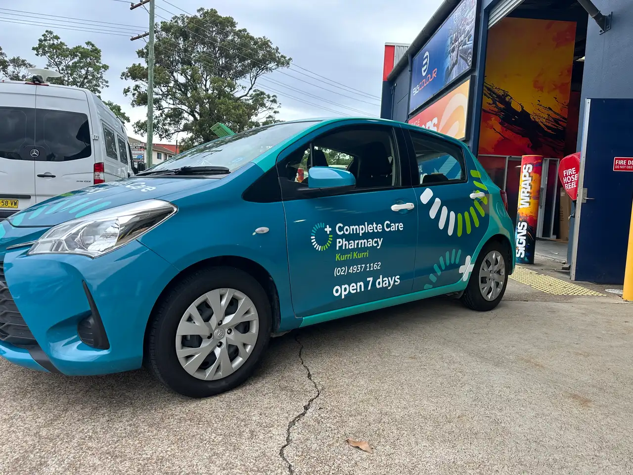 Driver's side of the turquoise wrapped Complete Care Pharmacy Yaris outside the Big Colour workshop, carrying the Kurri Kurri logo, (02) 4937 1162 and open 7 days