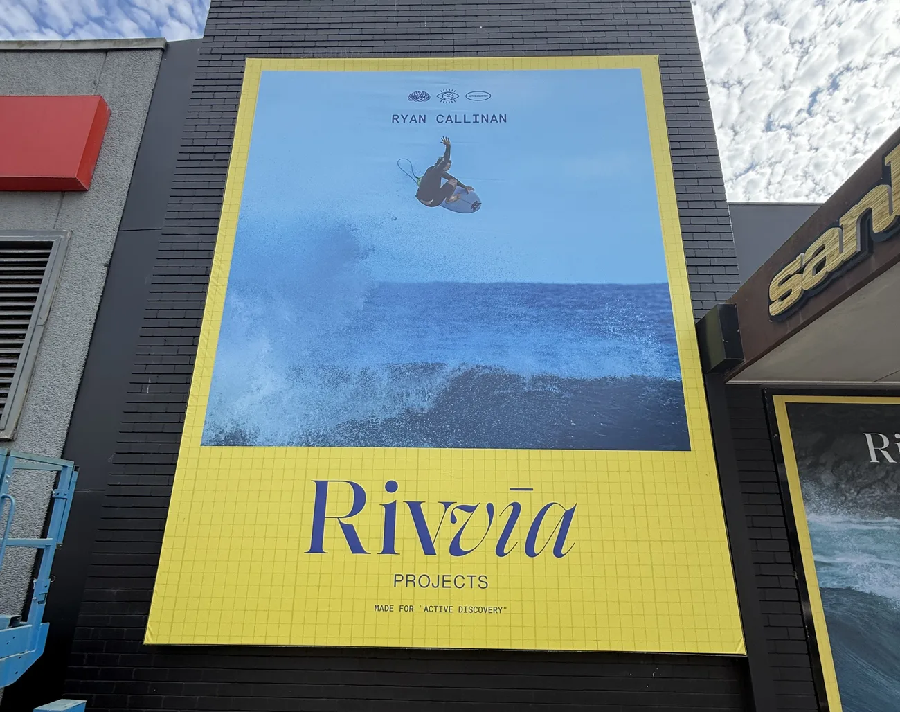 Large yellow Rivvia Projects banner sign with a surfing action photo, mounted on a black brick shopfront