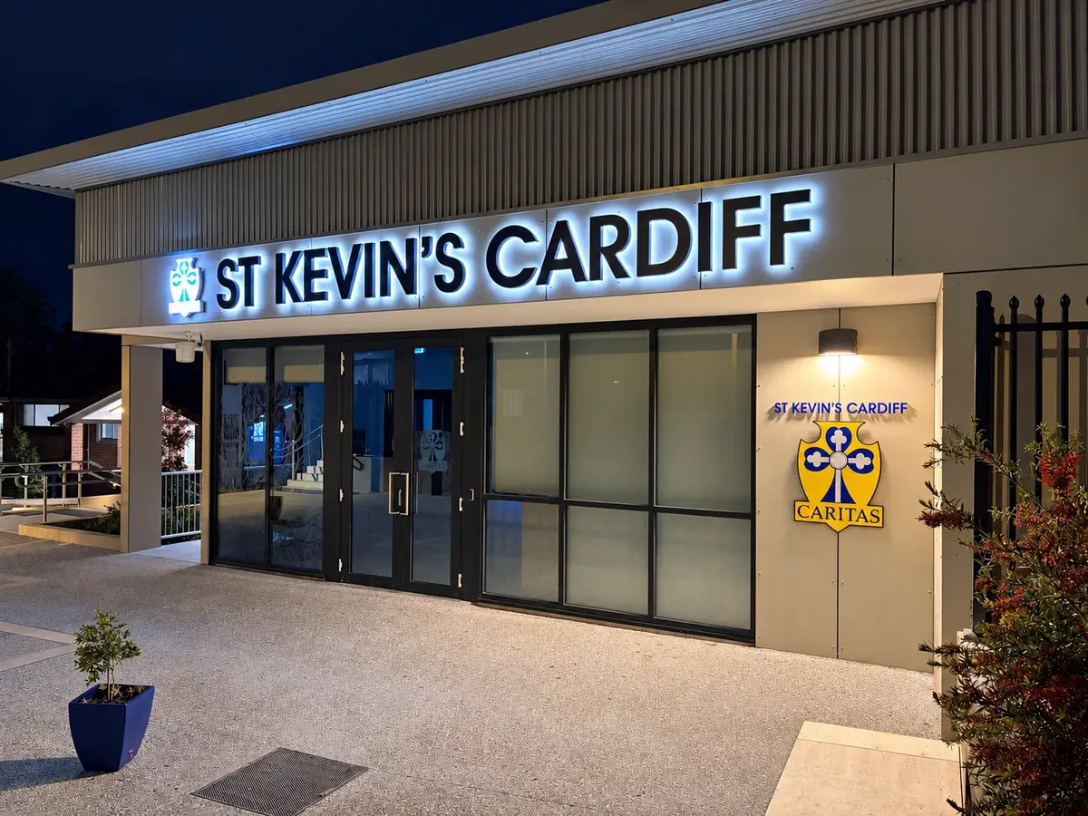 St Kevin's Cardiff illuminated sign above the glass entrance doors at night, with a Caritas crest sign on the wall