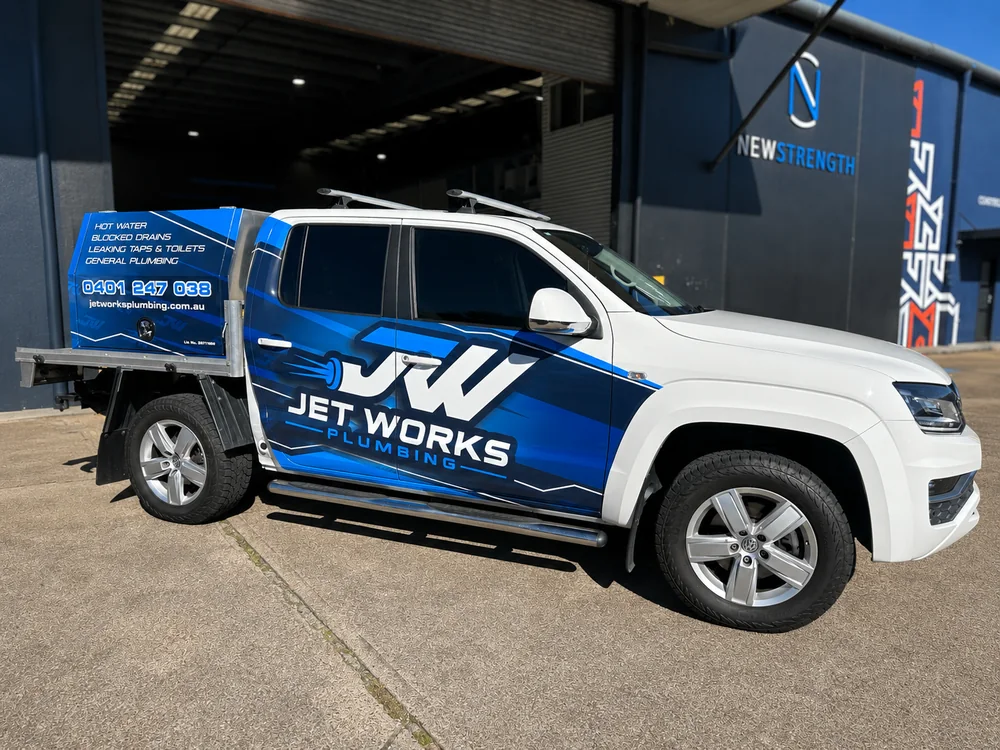 Jet Works Plumbing ute wrapped in white and blue company branding, with a toolbox on the tray, parked at commercial premises.