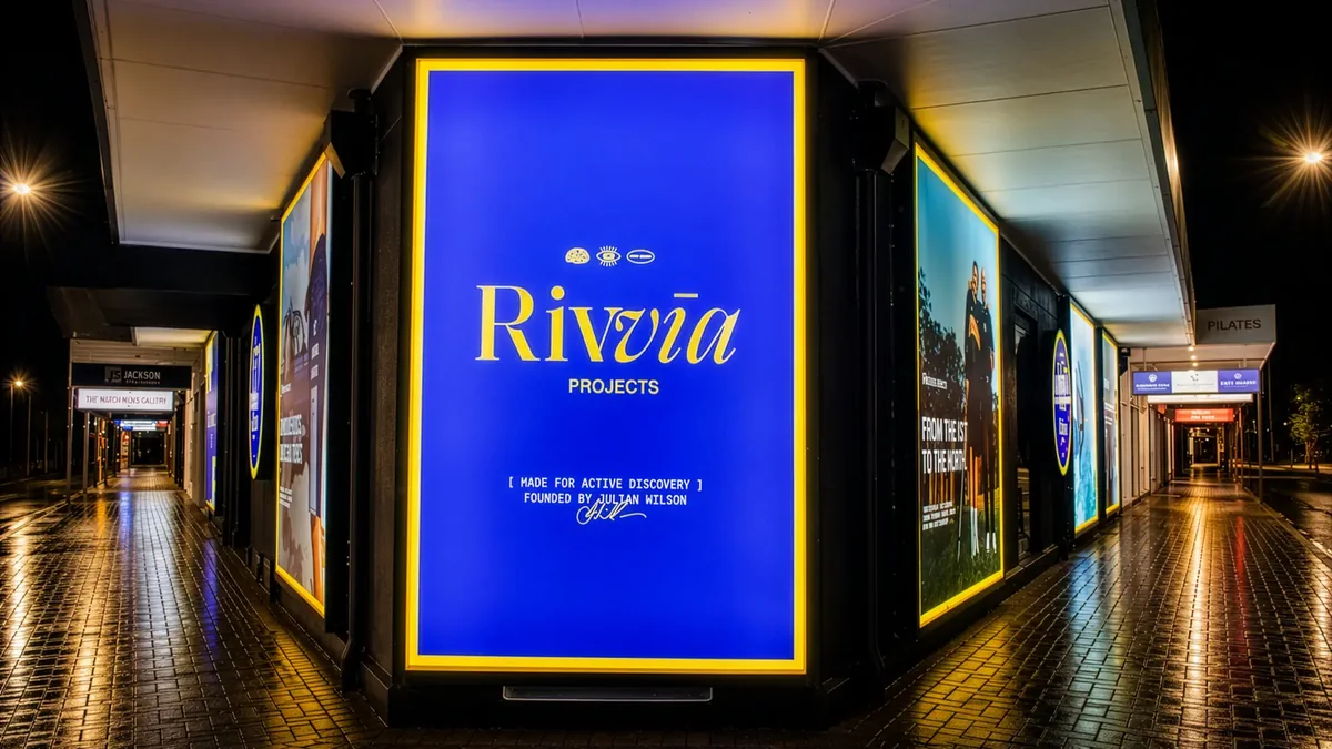 Illuminated shopfront lightbox signs wrapping a corner at night, glowing blue and yellow over the wet arcade paving
