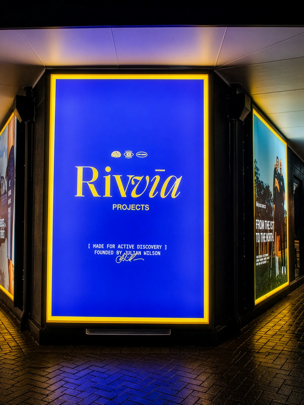 Rivvia Projects illuminated corner lightbox sign at night, blue face with a yellow-lit border